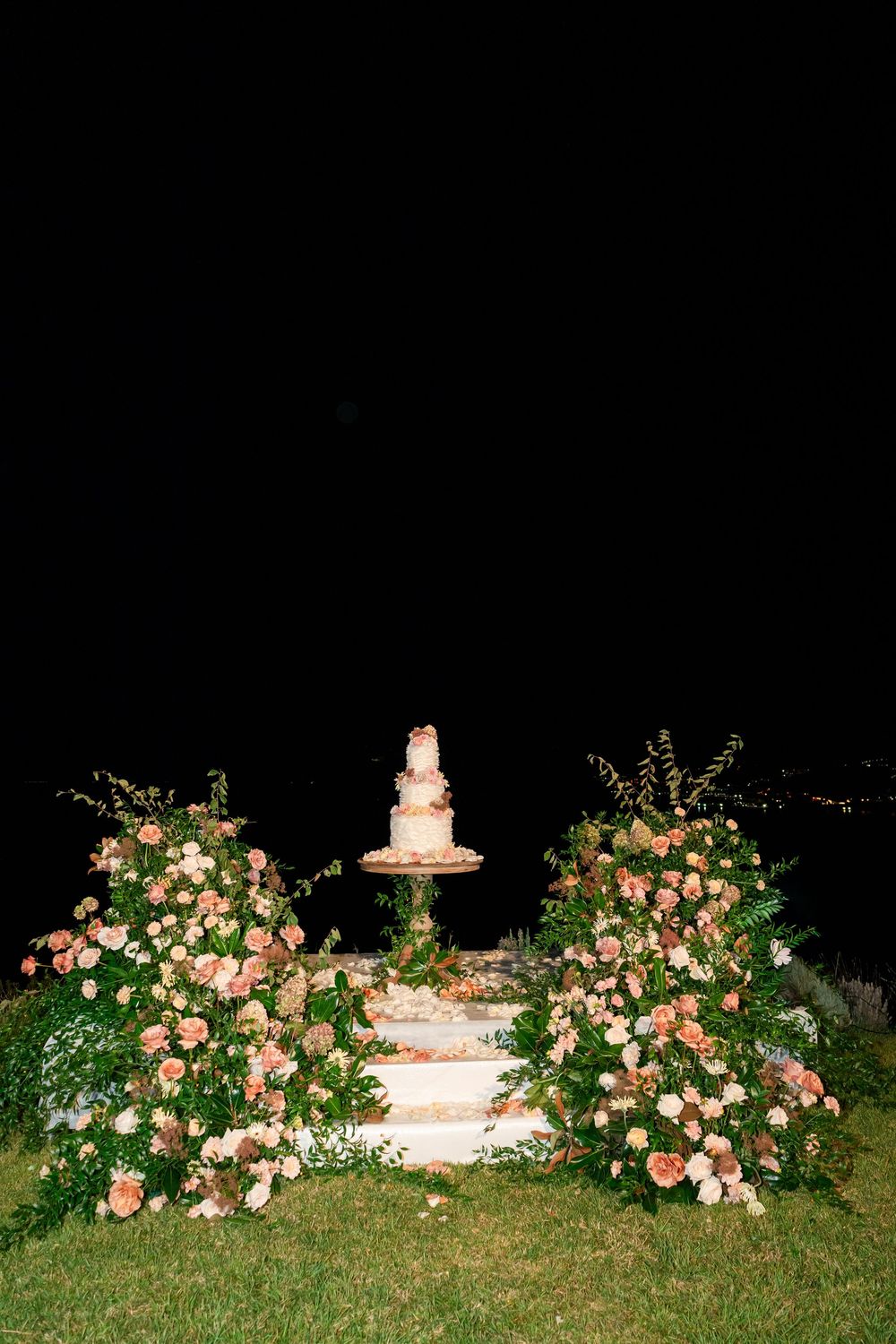 Wedding cake display decorated with cascading pink roses and greenery on a tiered stand at night.