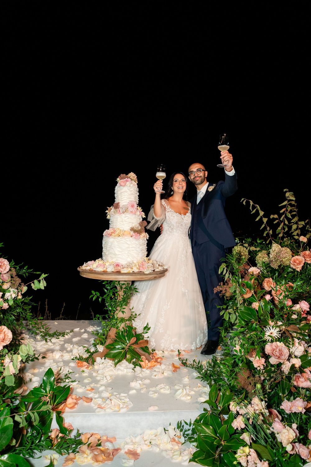 A wedding couple stands next to their elegant white tiered cake adorned with pink flowers and greenery at their reception.