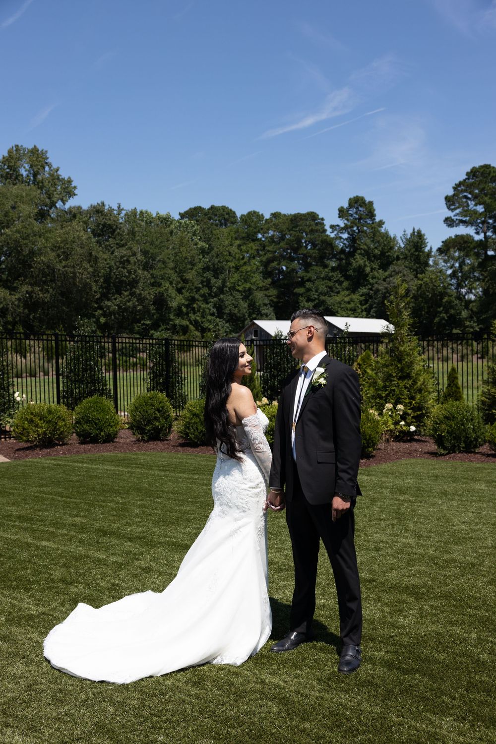 A couple in formal wedding attire shares a romantic moment on a lush green lawn with trees in the background.