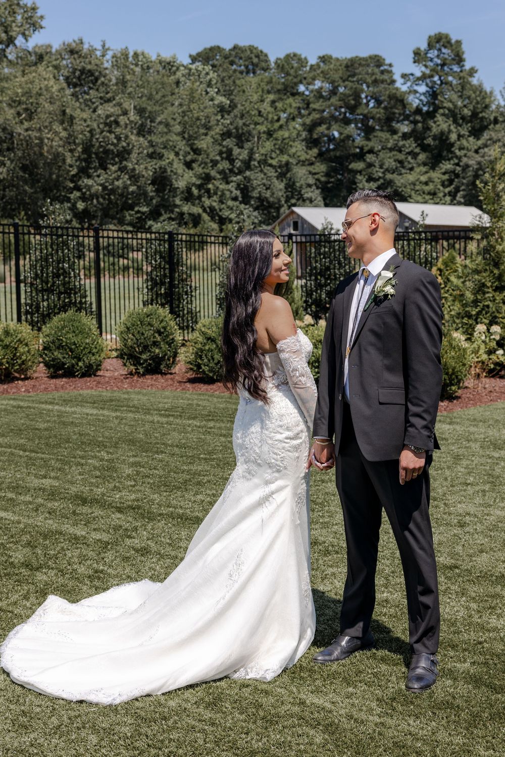 A wedding couple stands on manicured lawn with elegant landscaping and iron fencing in the background.
