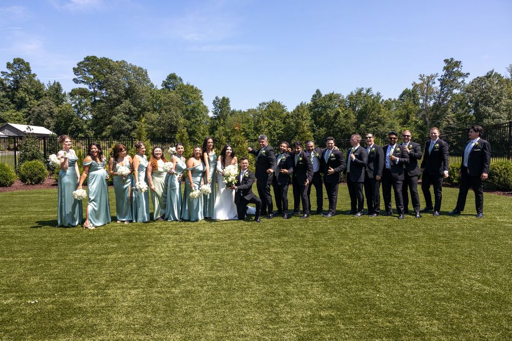 Wedding party members in formal attire stand in a line on green grass for group photos at an outdoor celebration.