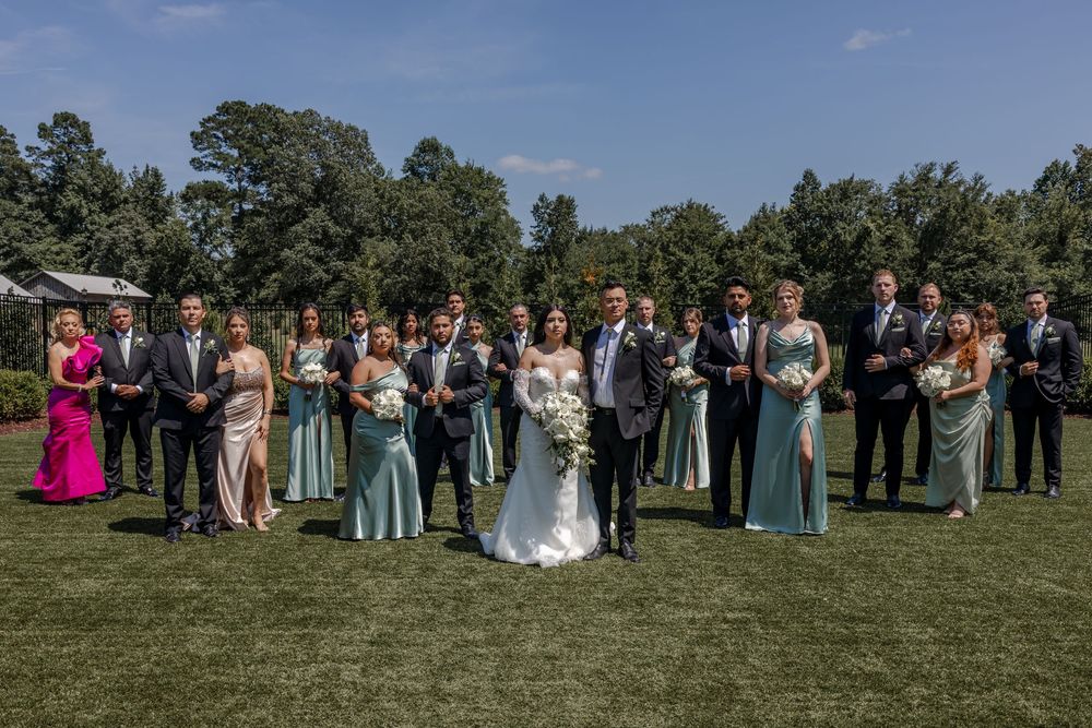 Wedding party members stand in line on a grassy lawn for formal group photos in varying shades of blue attire.