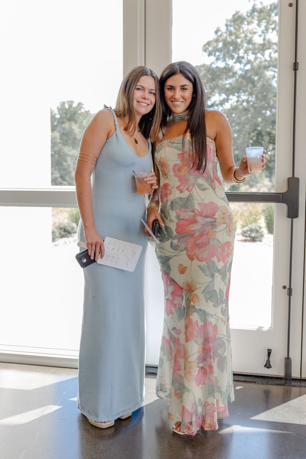 Two women in light blue and floral dresses stand together smiling at an indoor event.