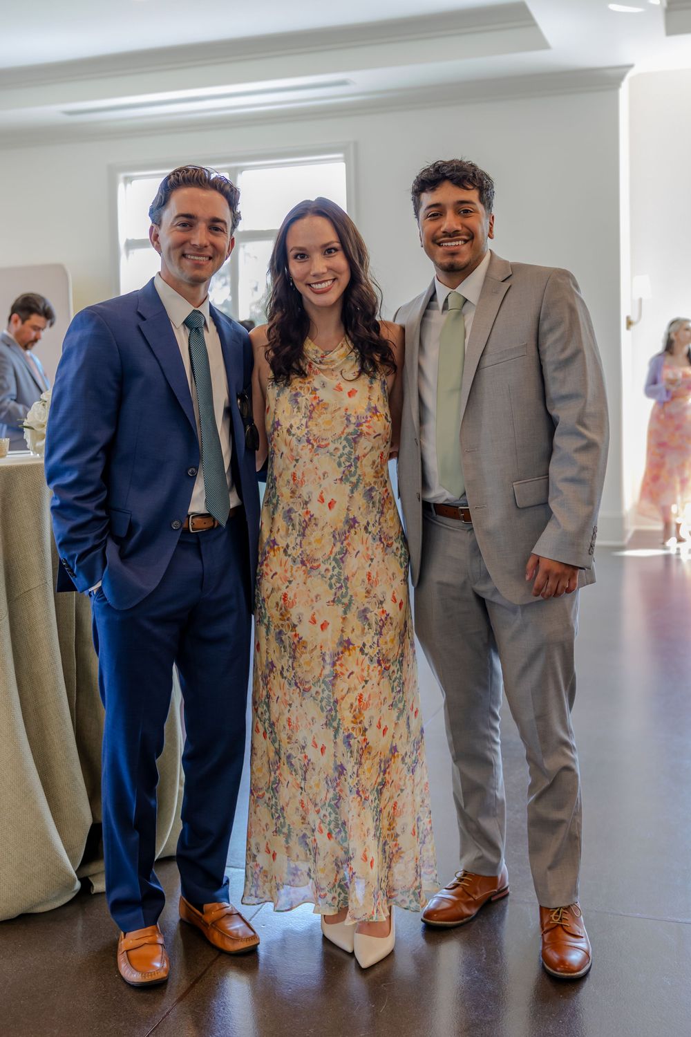 Three people in formal attire stand together at an event, with one wearing a floral dress and others in suits.