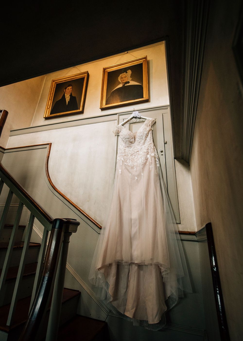 White wedding dress hanging in a dimly lit stairwell with vintage portraits on the wall.