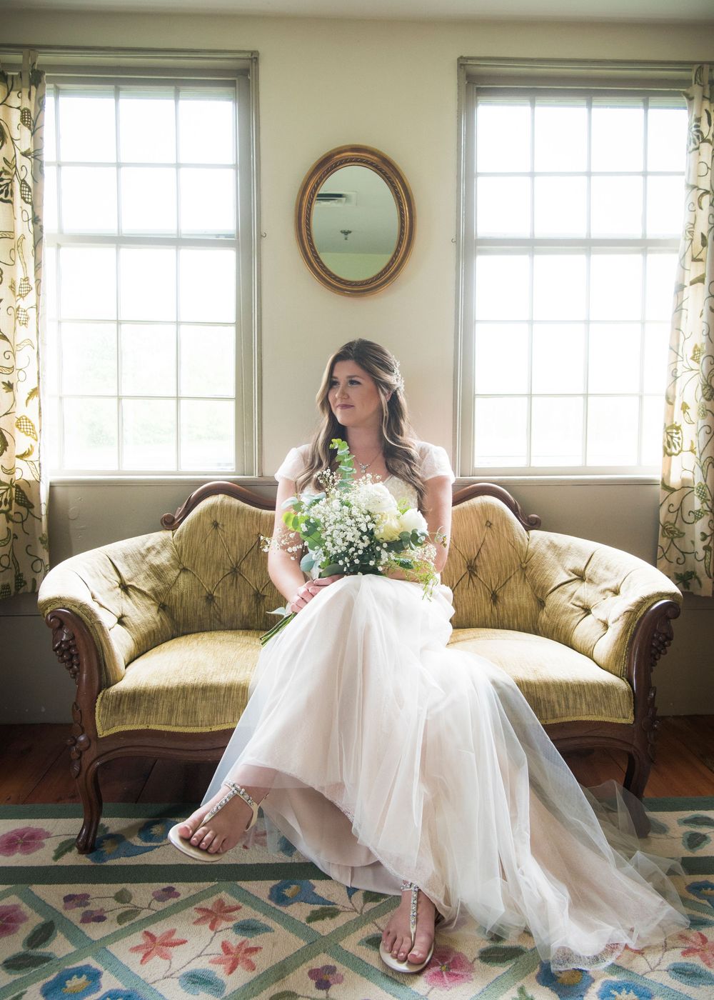 White wedding dress displayed on vintage floral sofa near bright windows.