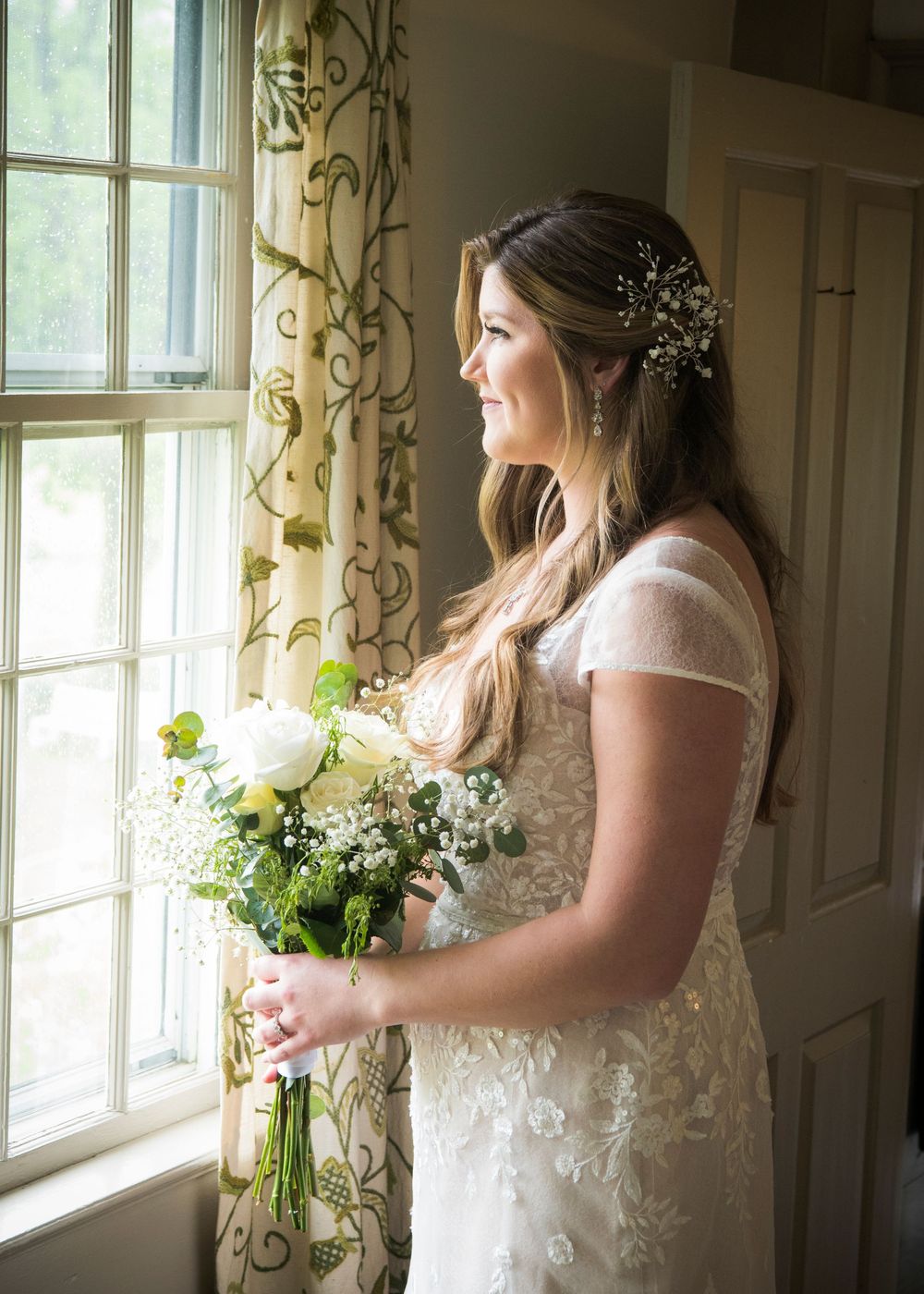 Bride in a white lace dress stands pensively by a sunlit window holding a bouquet of flowers.