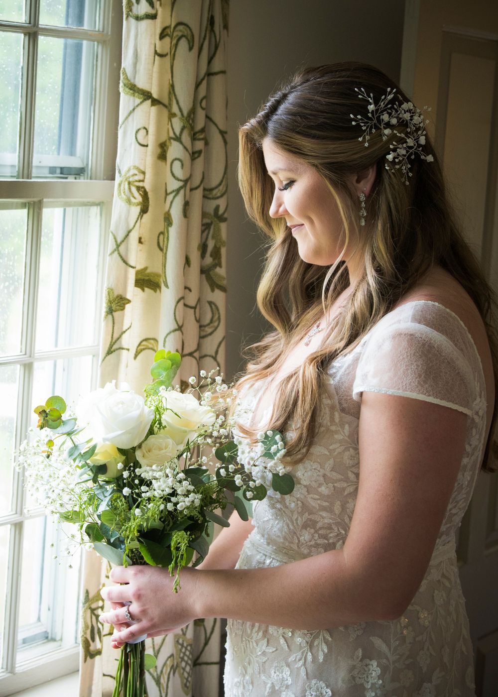 Bride in lace dress stands by sunlit window holding delicate floral bouquet with greenery.