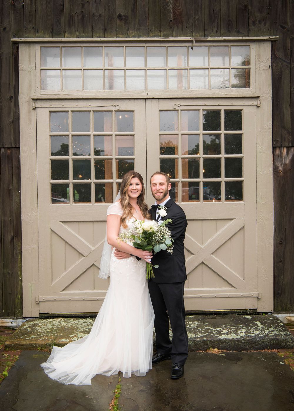 Newlyweds pose in front of a vintage barn door holding a white floral bouquet.