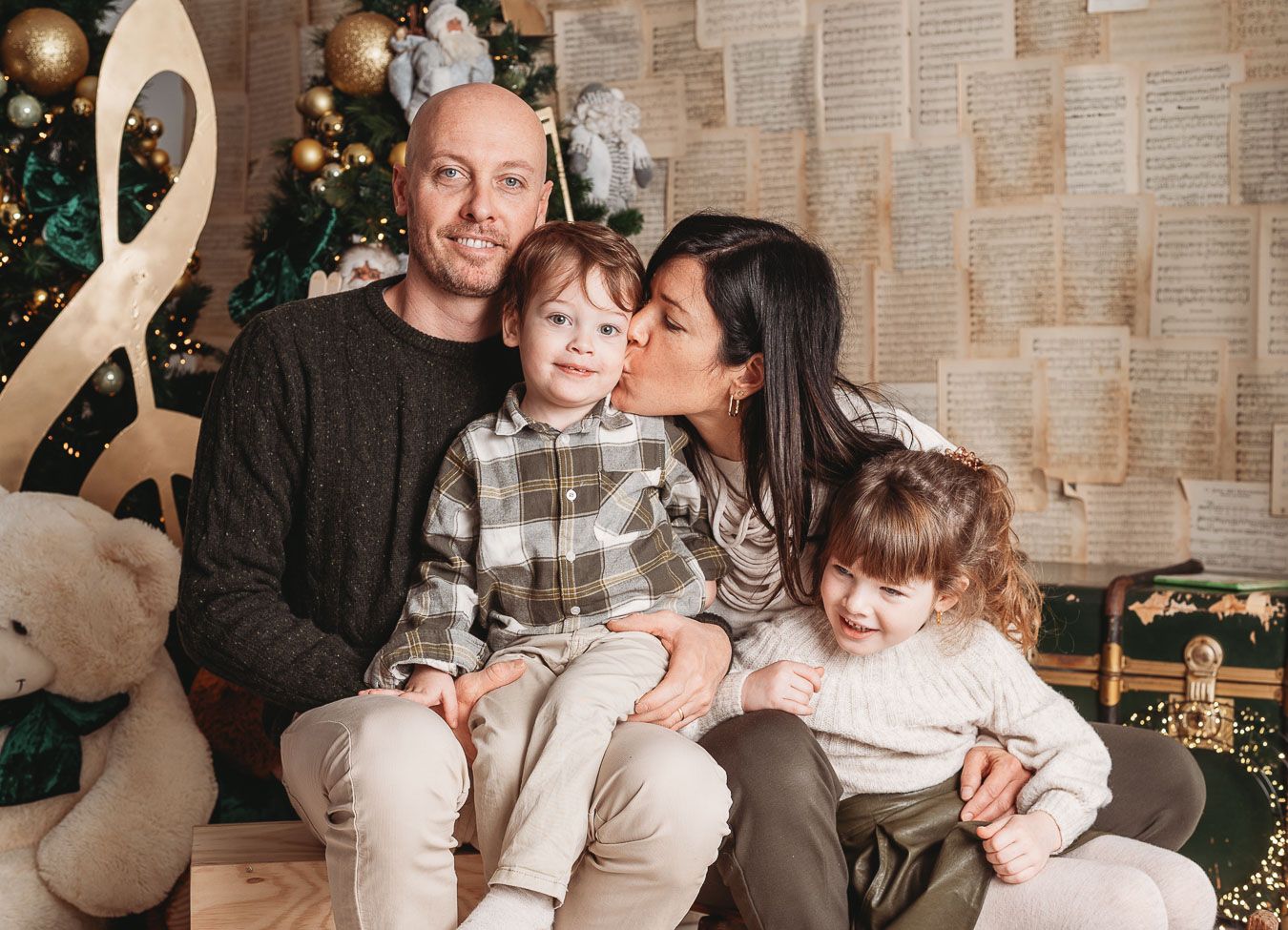 Family wearing matching sweaters sits together on couch during holiday season with Christmas tree in background.