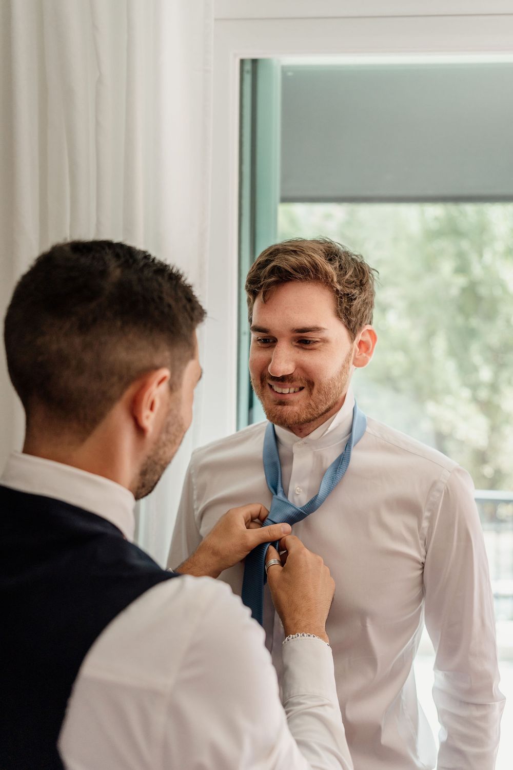 Someone helps adjust a blue necktie on a person wearing a white dress shirt near a window.
