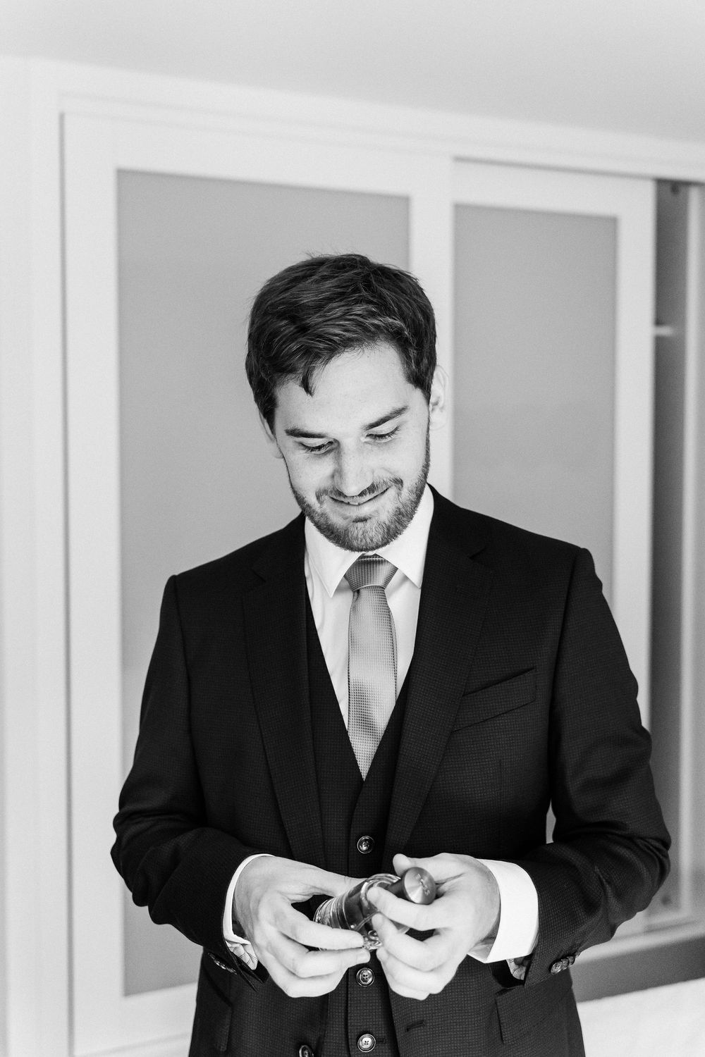 A smiling person in a black suit and tie looks down at something in their hands in a black and white photo.