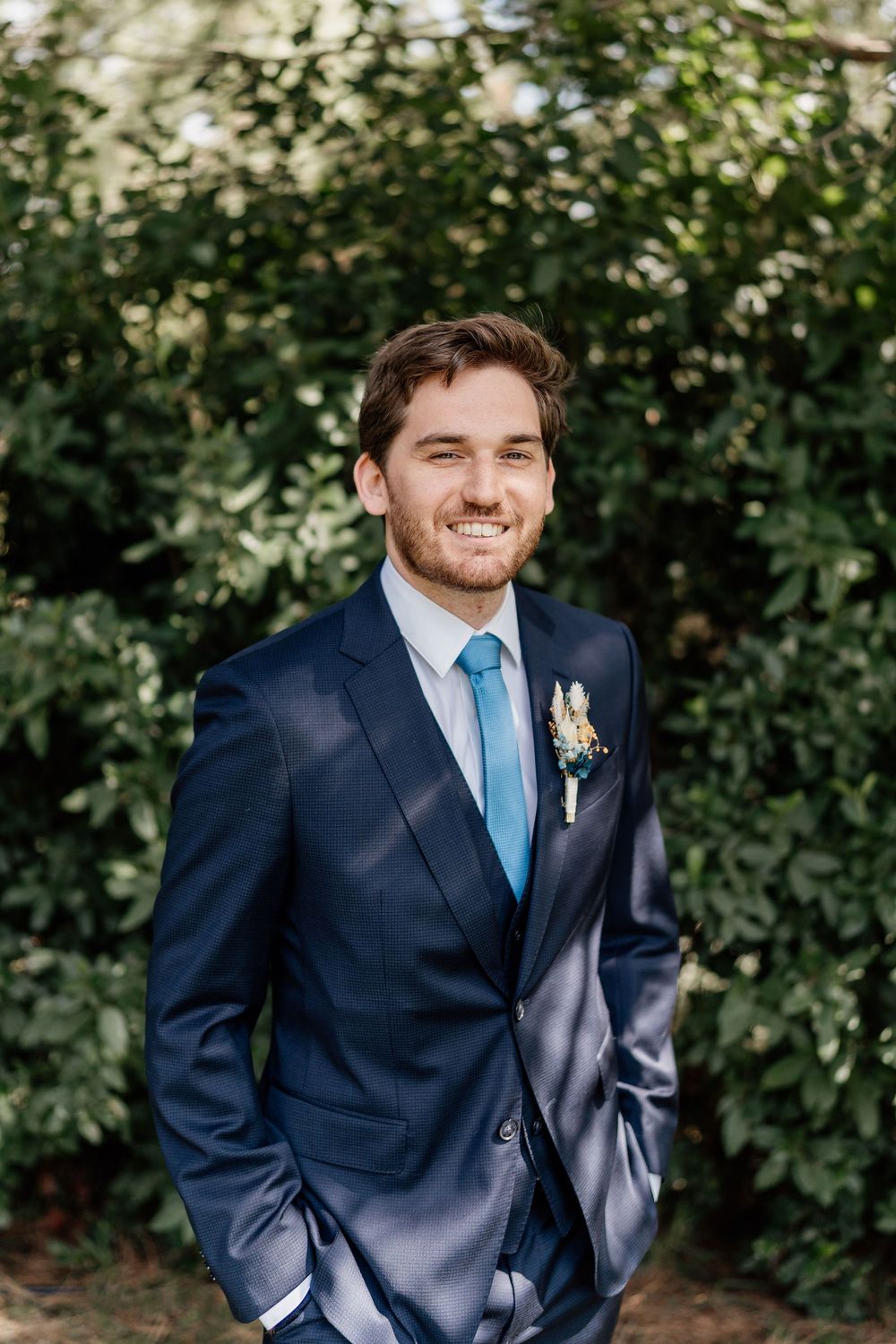 A groom in a navy suit and blue tie poses outdoors against a leafy green background.