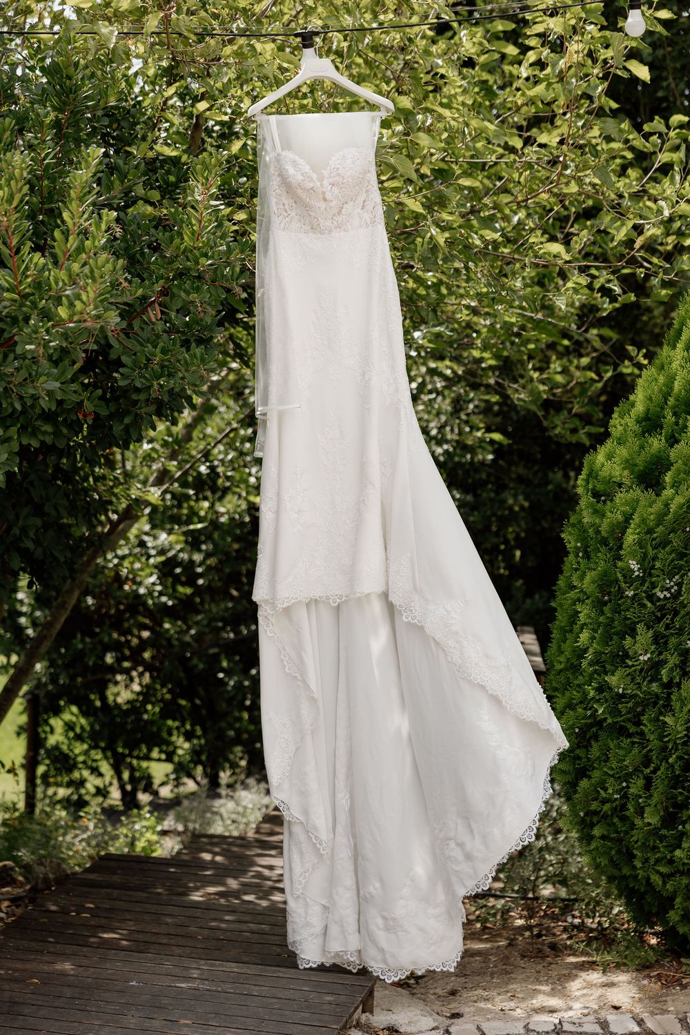An elegant white wedding dress hangs on a wooden hanger against a backdrop of green foliage in a garden setting.