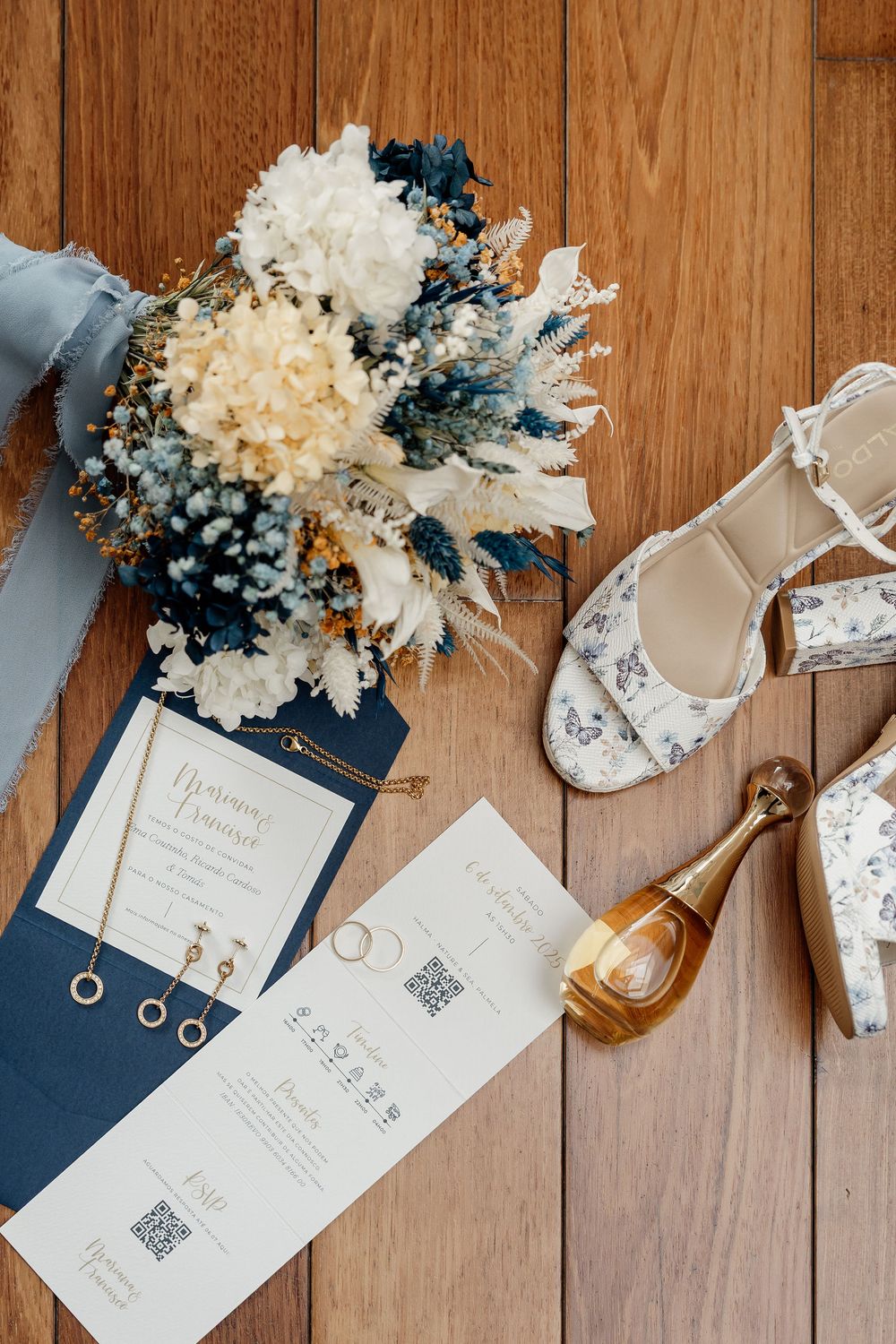 Wedding details flatlay with dried flower bouquet, navy blue invitations, floral heels and champagne bottle on wooden floor.