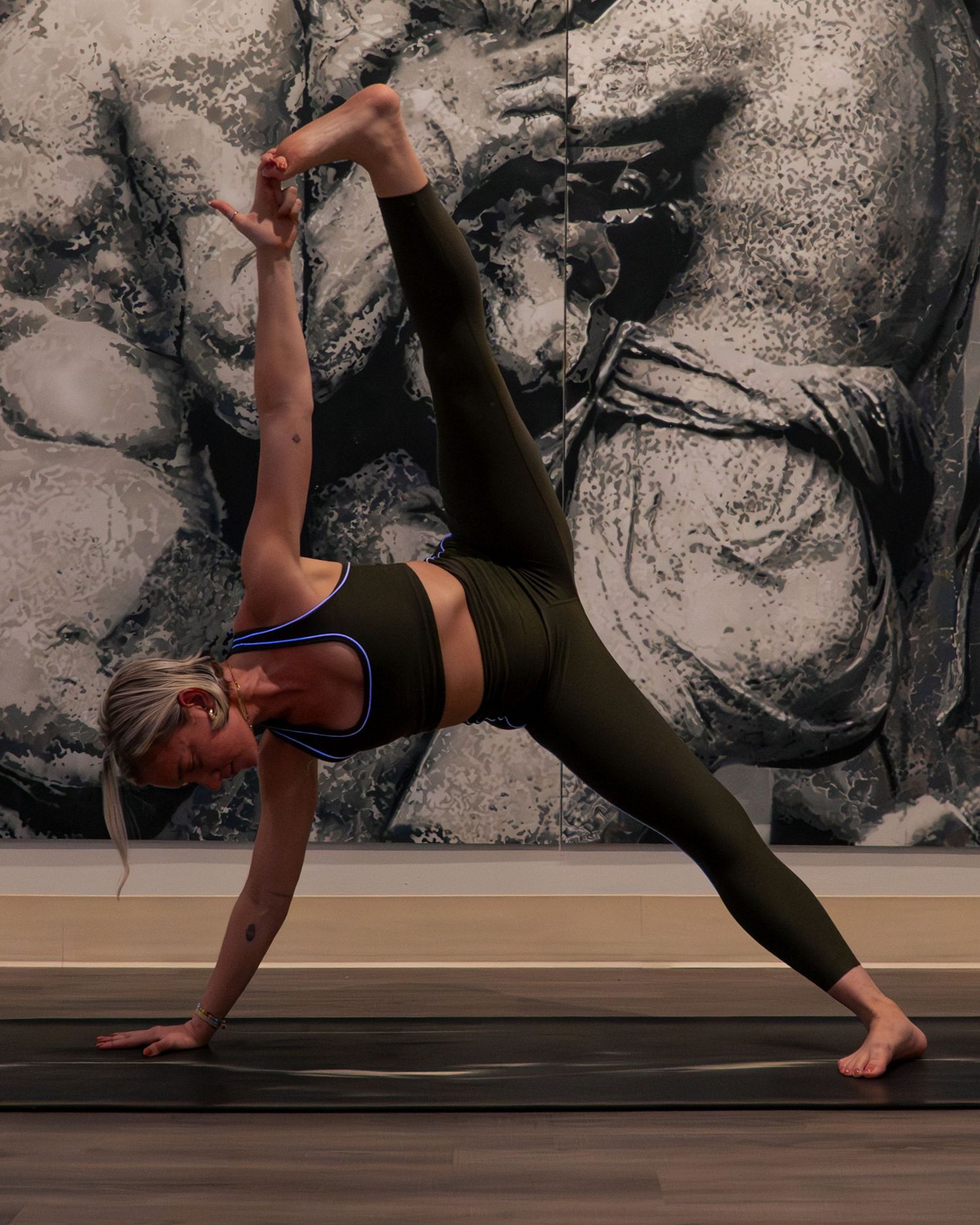 A person performs a side plank yoga pose on a yoga mat against a black and white abstract wall backdrop.