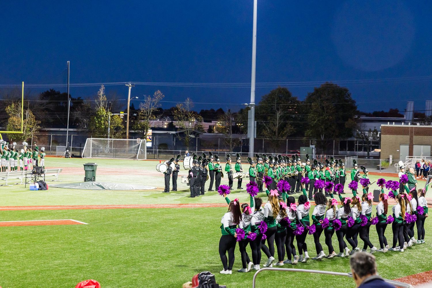 High school marching band members perform in formation on a sunny football field during an outdoor practice session.