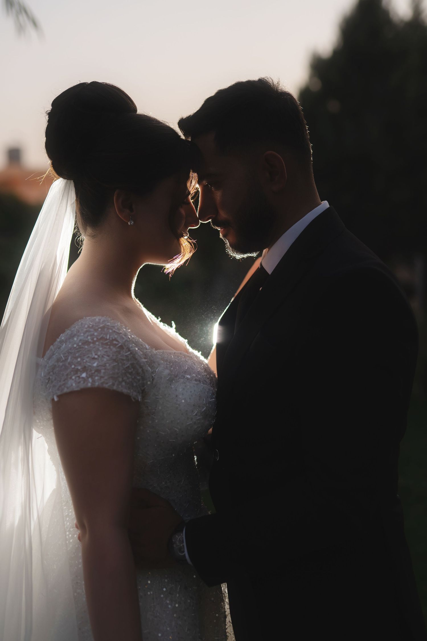 Silhouetted couple in wedding attire share an intimate moment at sunset, with bride's veil flowing in backlit scene.