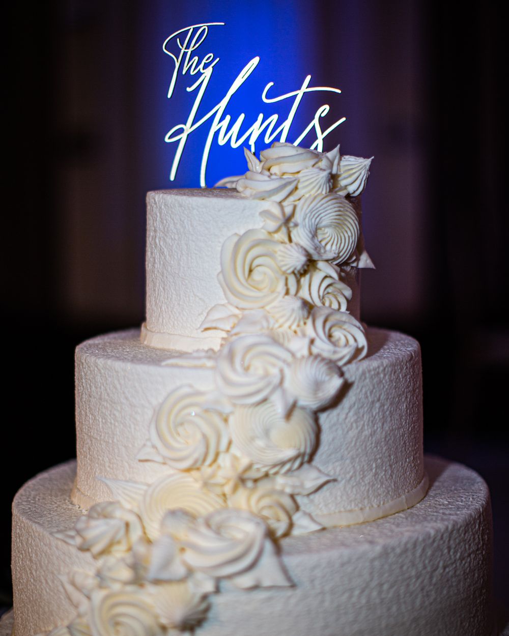 Elegant white wedding cake with cascading floral decorations illuminated by blue lighting.