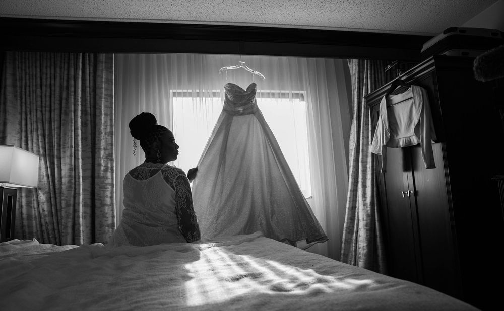 Wedding dress hanging in silhouette against window light in hotel room.