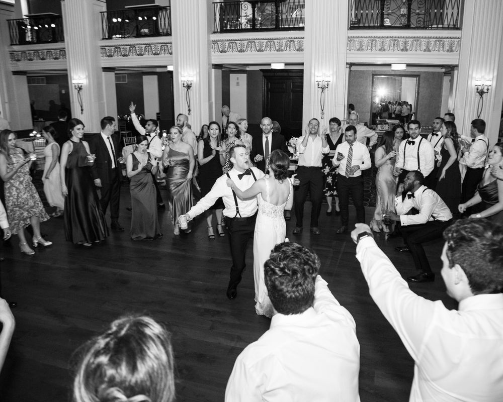 Wedding guests dance and celebrate in an elegant ballroom with ornate architectural details.