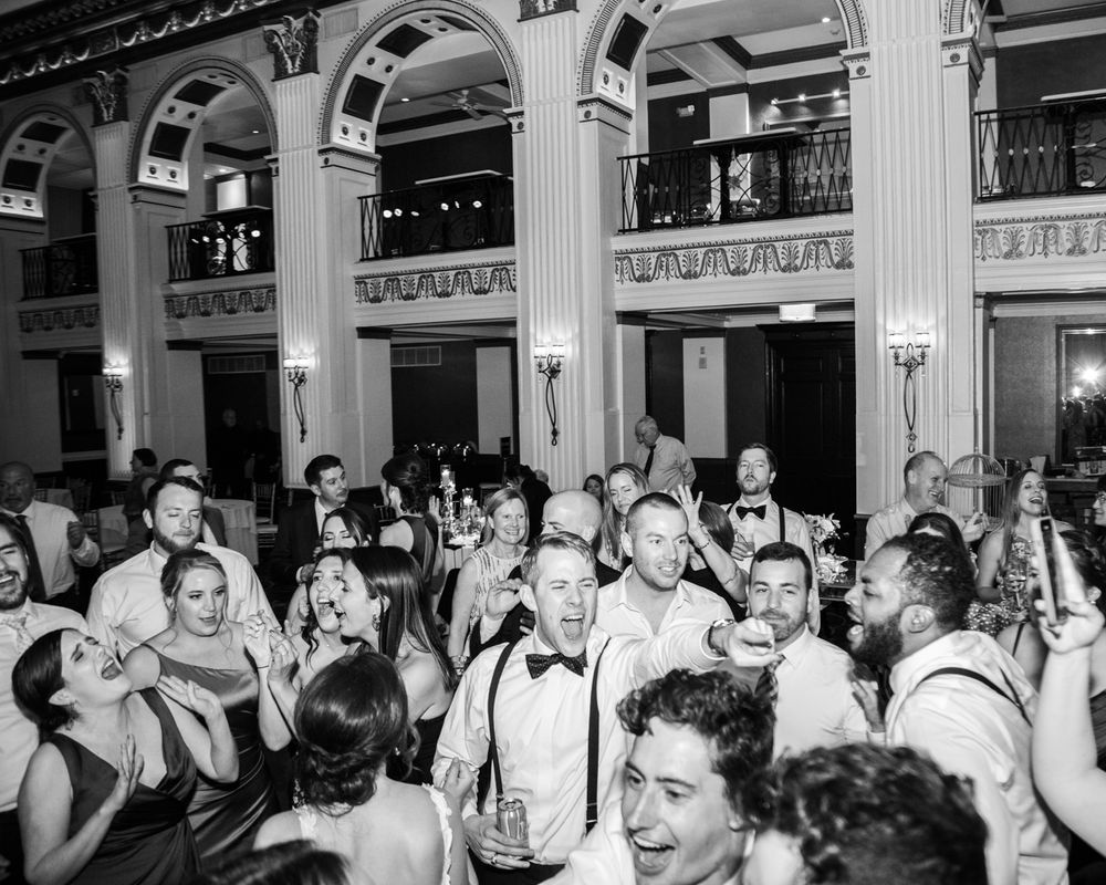 Large group of wedding guests dancing and celebrating in a grand ballroom with classical columns.