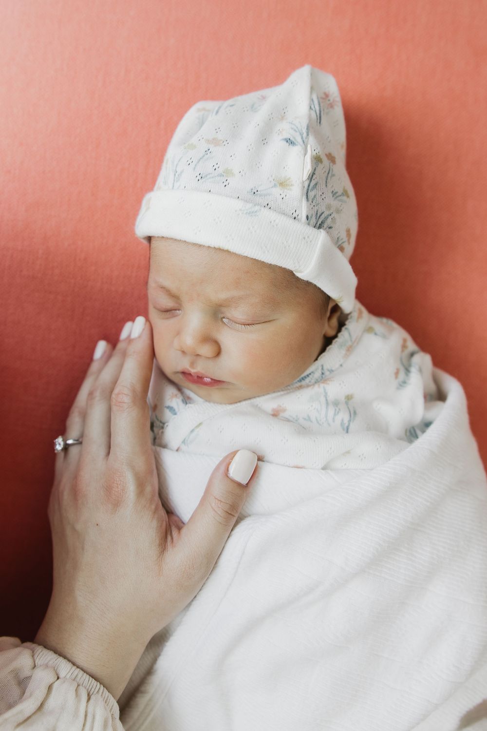 A sleeping newborn baby wrapped in white swaddling cloth and wearing a soft cap against a coral-colored background.