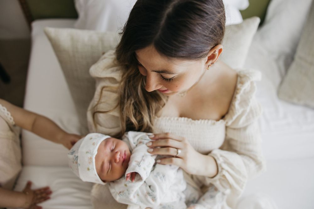 A mother in white dress tenderly embraces and nurses her newborn baby in a bright, intimate setting.