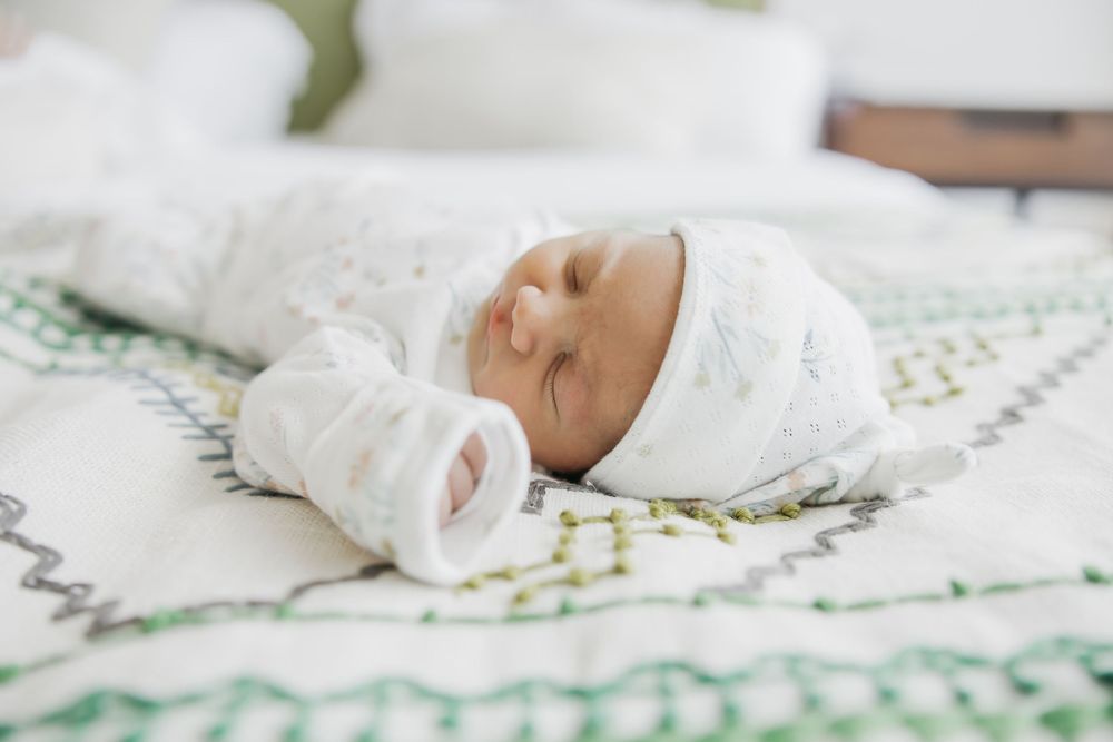 A sleeping newborn baby swaddled in white blankets rests peacefully on a patterned green and white blanket.