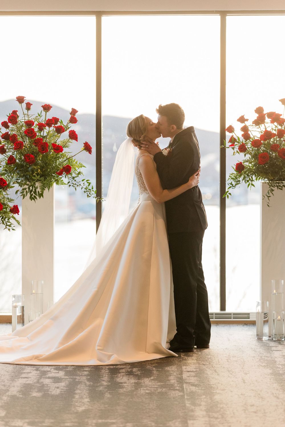 A romantic wedding ceremony silhouette with elegant red rose arrangements flanking the couple against large windows.