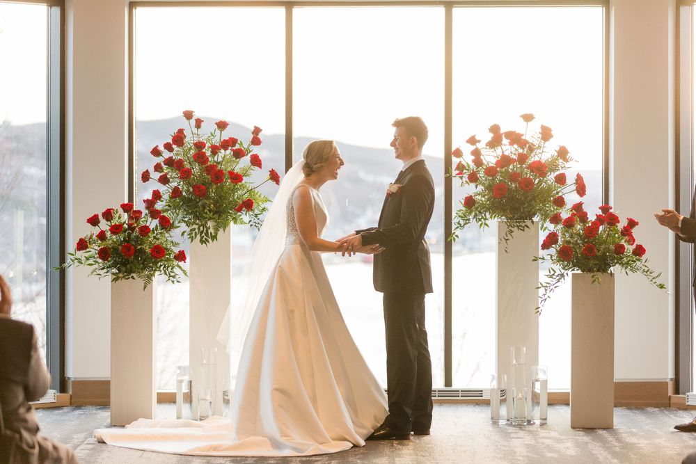 A wedding couple stands in front of large windows with red floral arrangements creating a romantic silhouette.