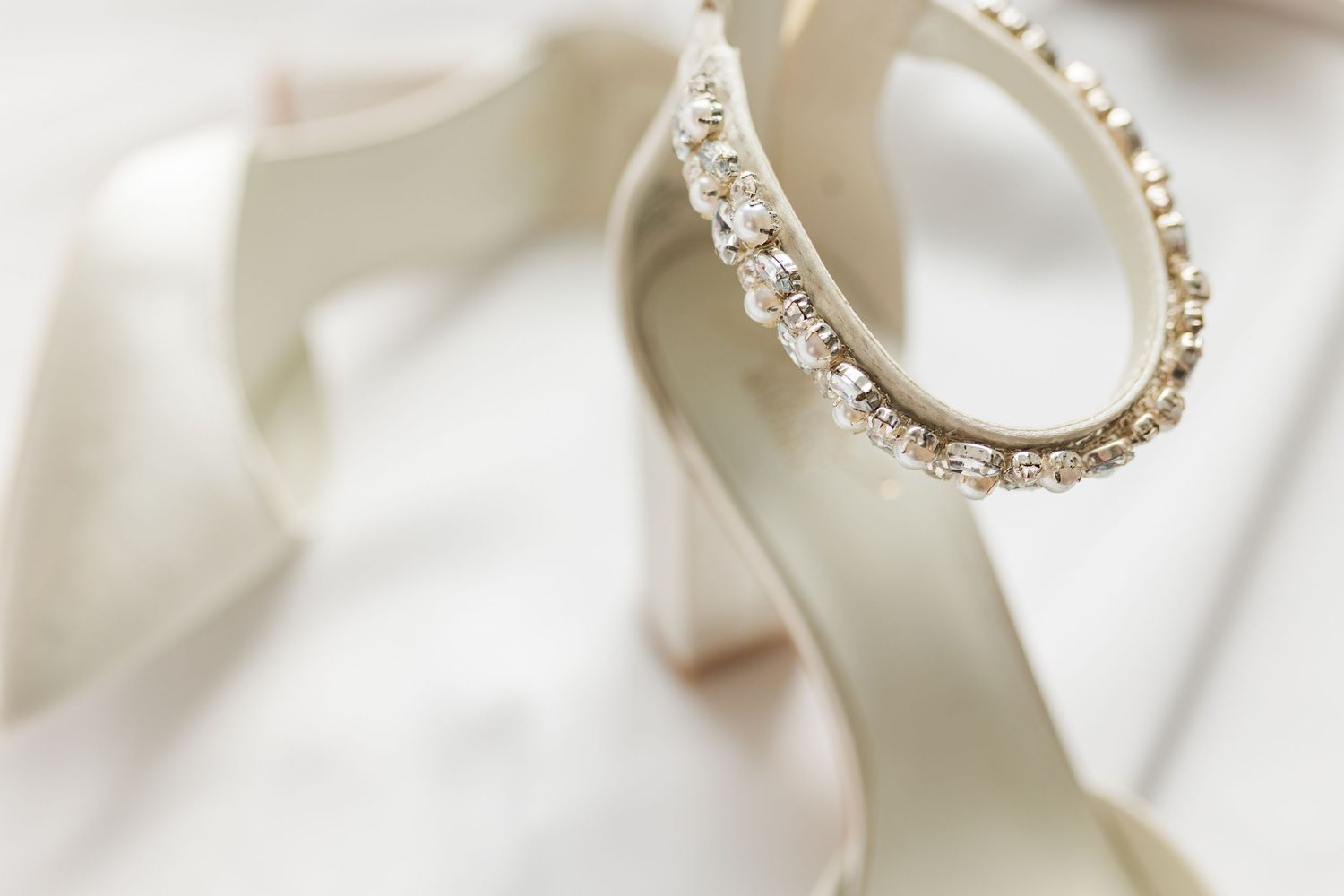 Close-up detail of crystal-adorned white wedding shoes against soft white textile backdrop.