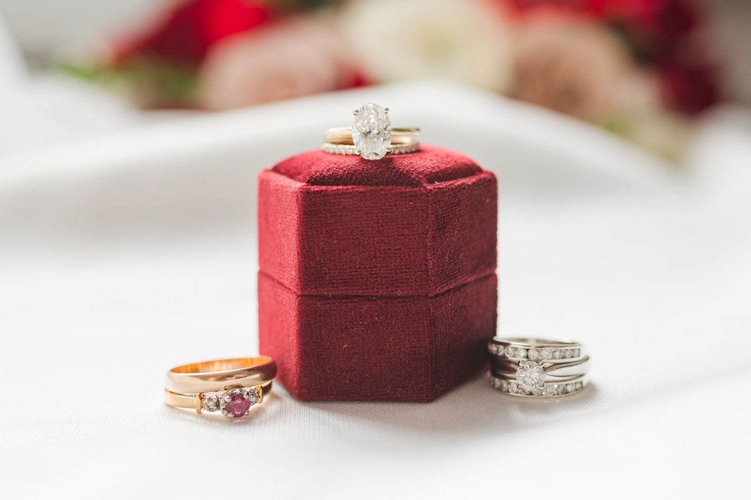 Diamond engagement ring sits atop red velvet hexagonal ring box with two gold bands beside it.