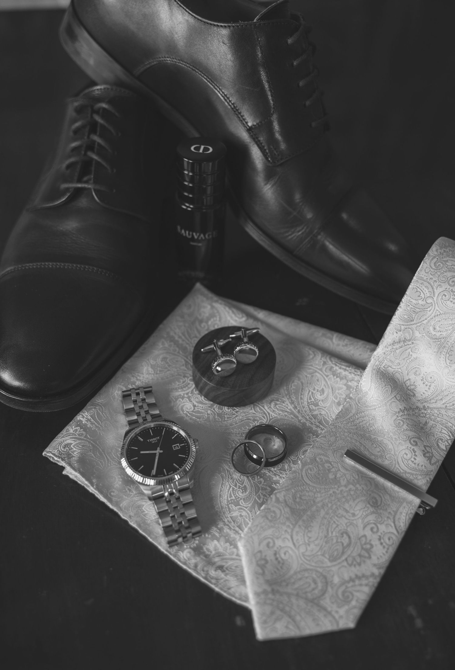 Black dress shoes and accessories including watches arranged on decorative patterned fabric.