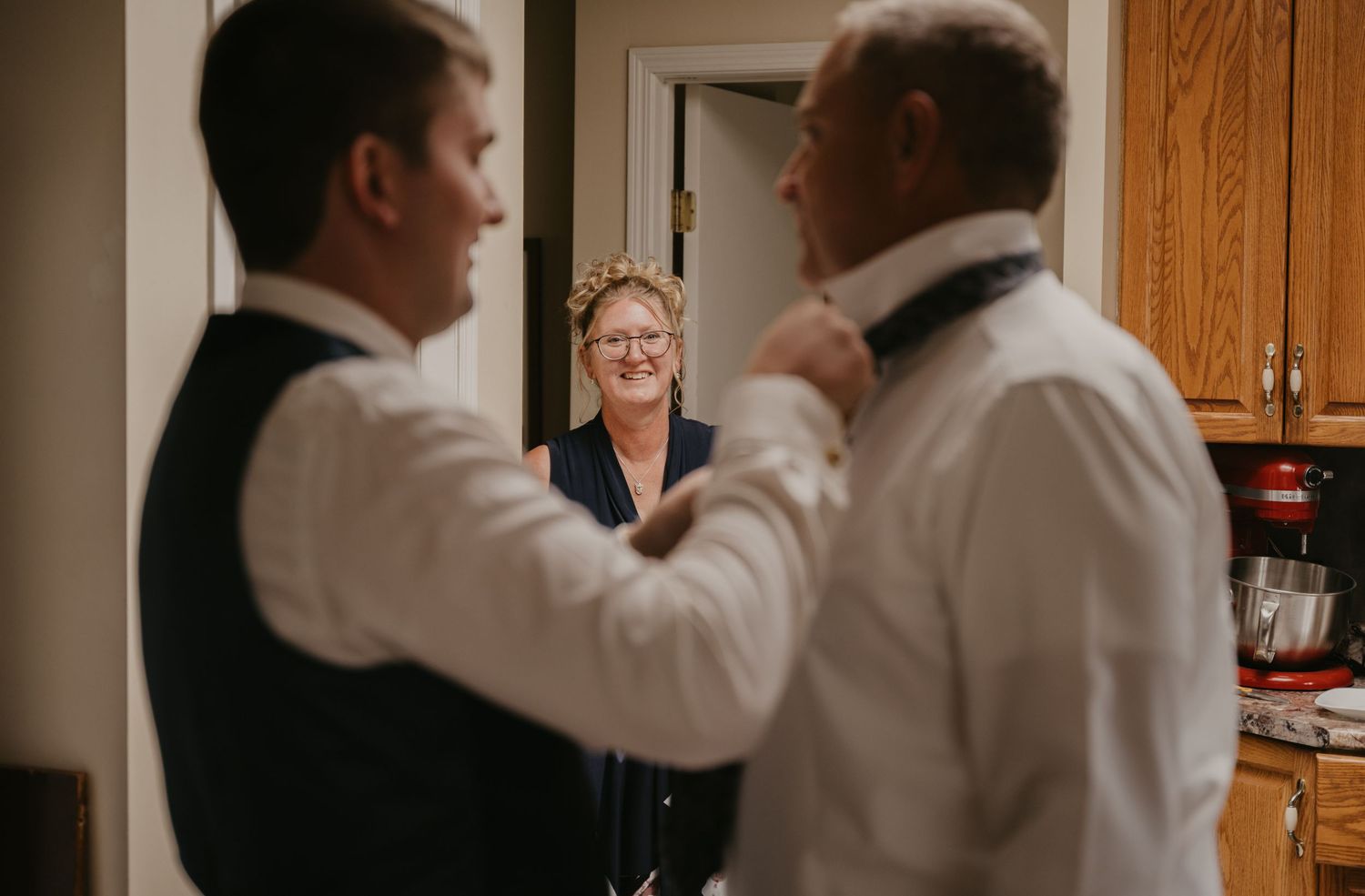 Wedding preparation scene showing people helping with formal attire in a kitchen setting.