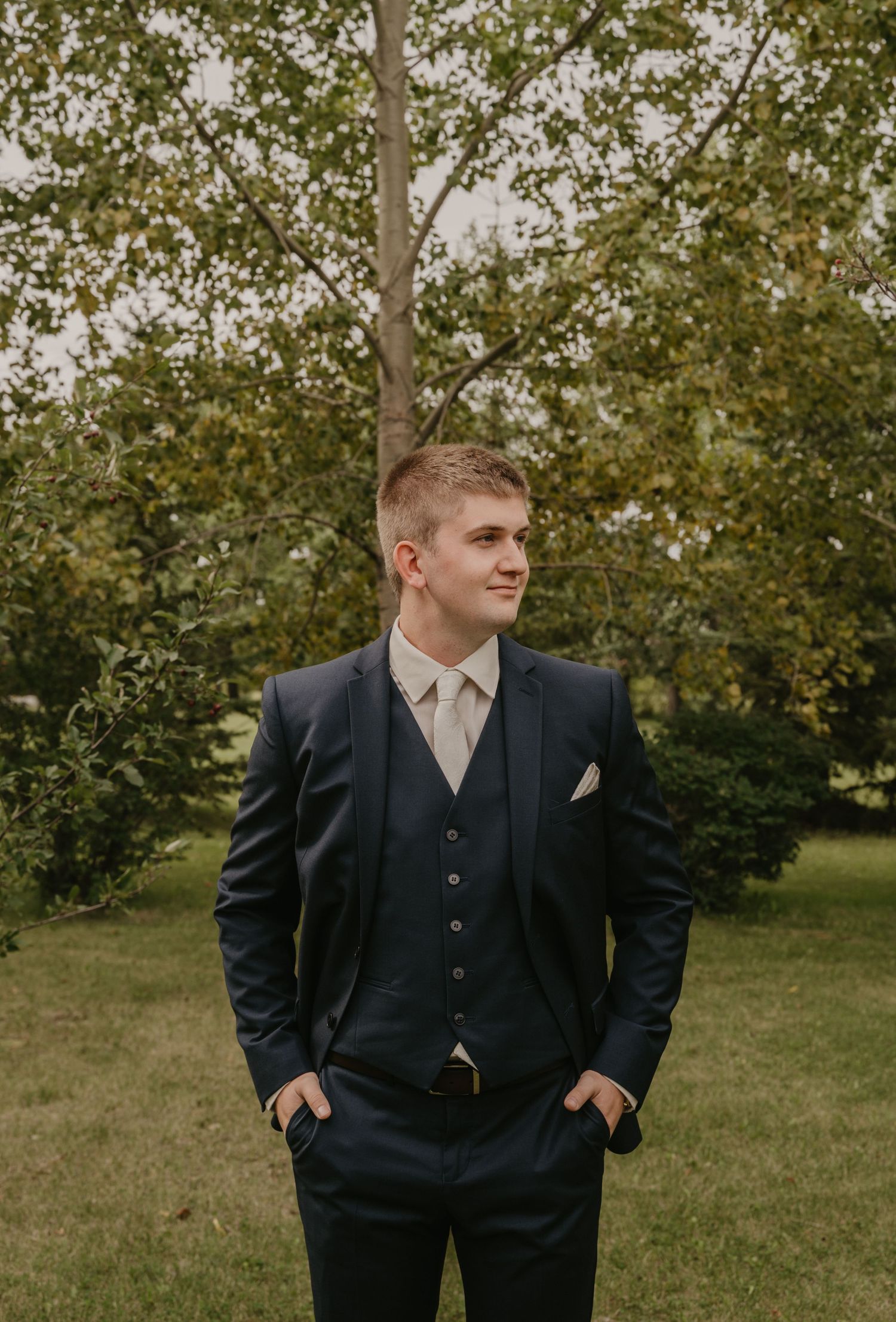 Groom in a classic black suit poses outdoors in front of autumn trees.