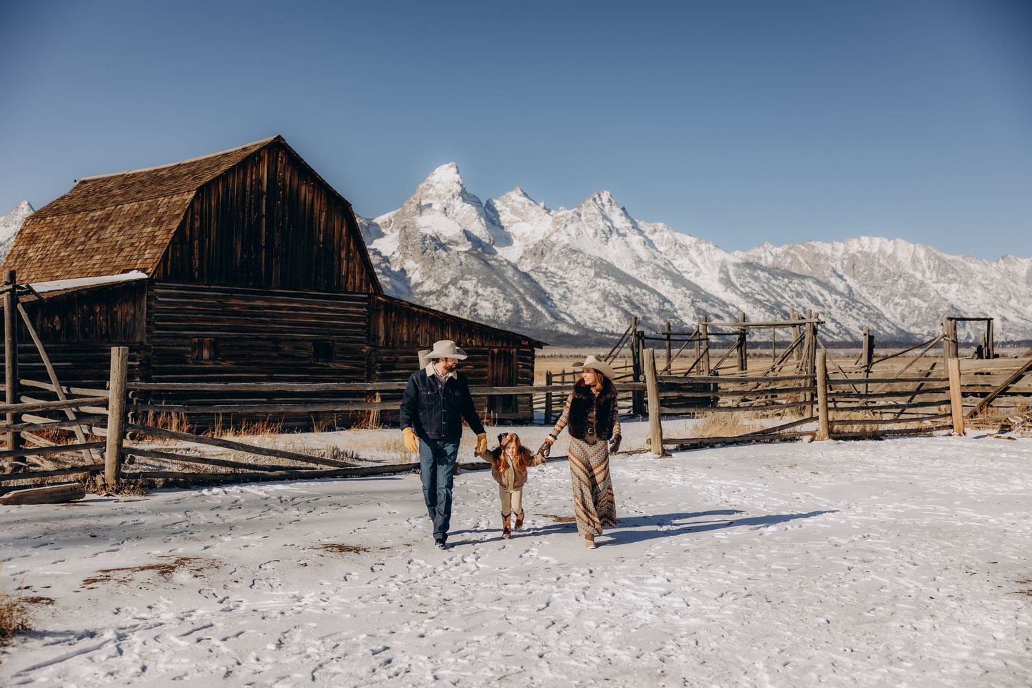 A panoramic view of snow-covered mountains with rustic wooden barns and fences in Grand Teton National Park.