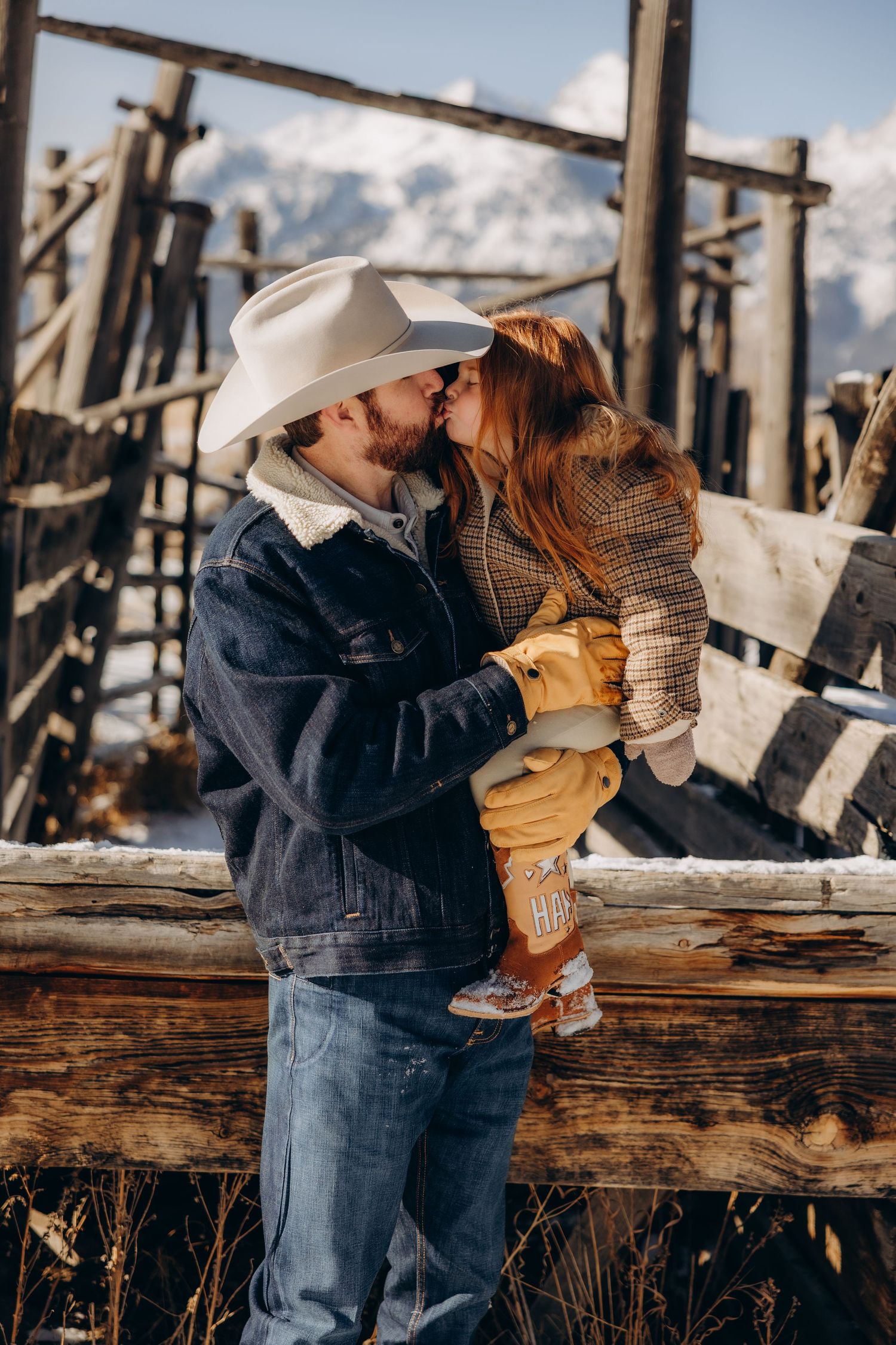 A couple shares an intimate moment against a rustic wooden fence with snowy mountains in the background.