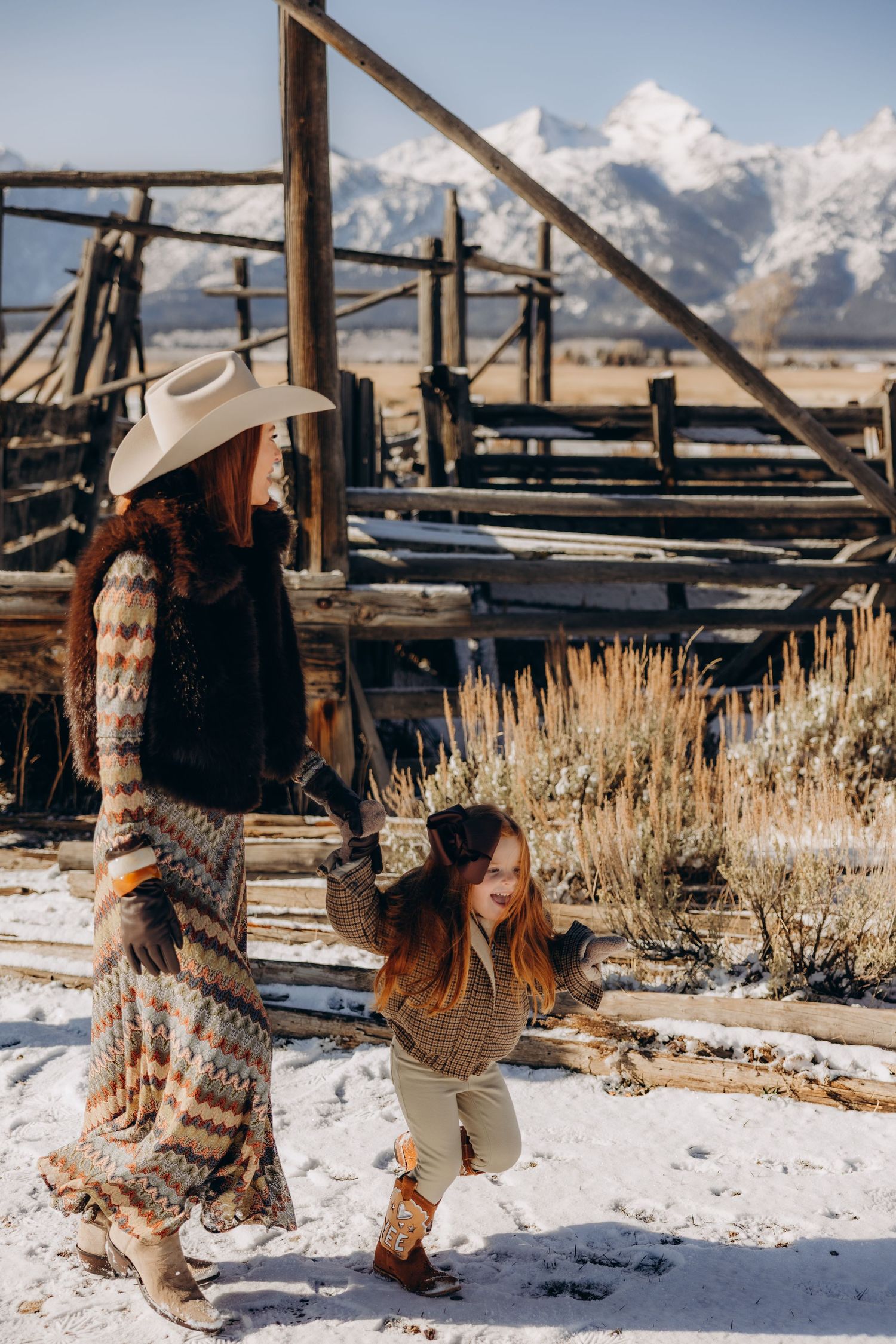 A couple dances playfully in the snow with rustic wooden structures and mountain peaks behind them.