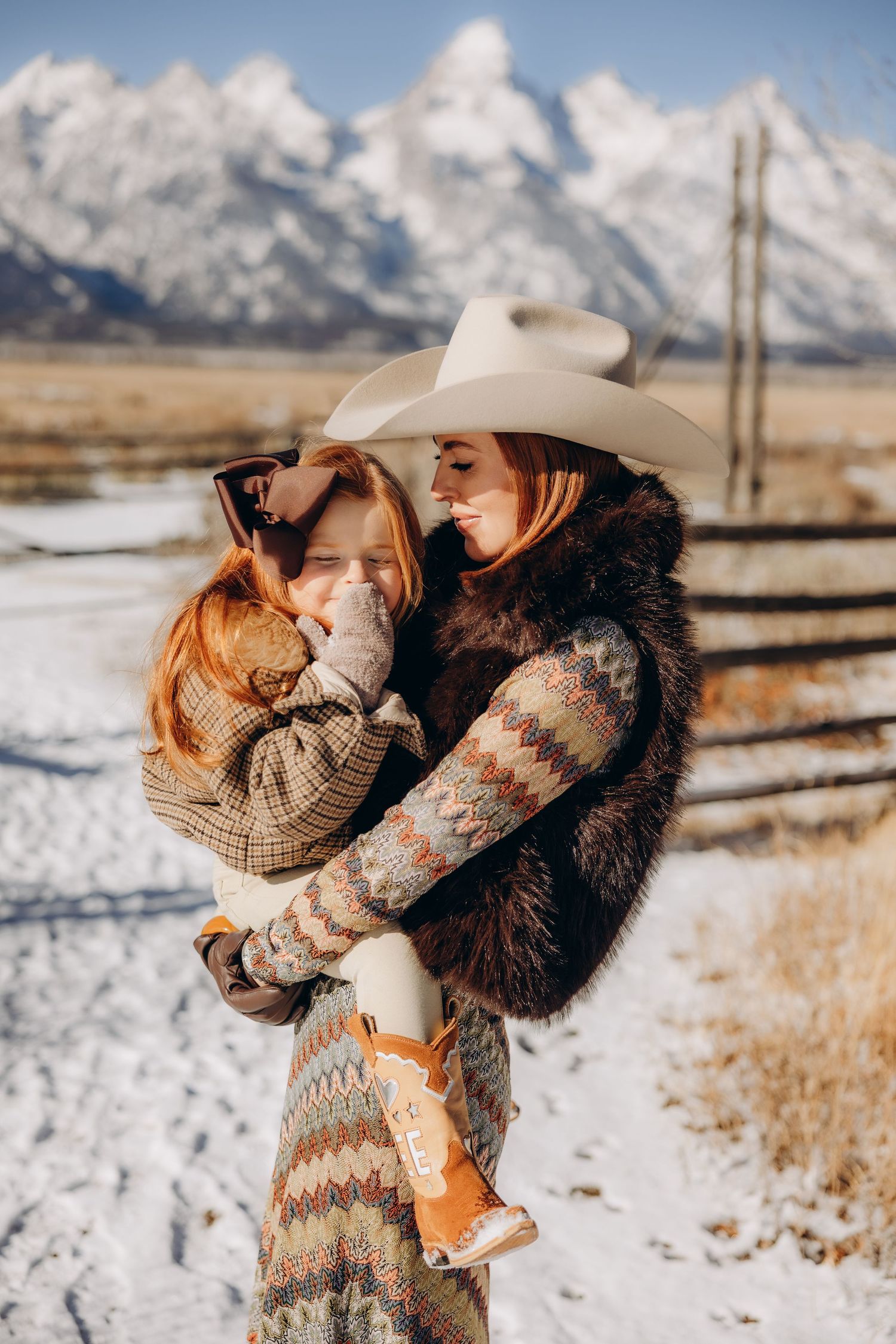 A couple embraces in a snowy winter landscape with mountains and wooden fences in the background.