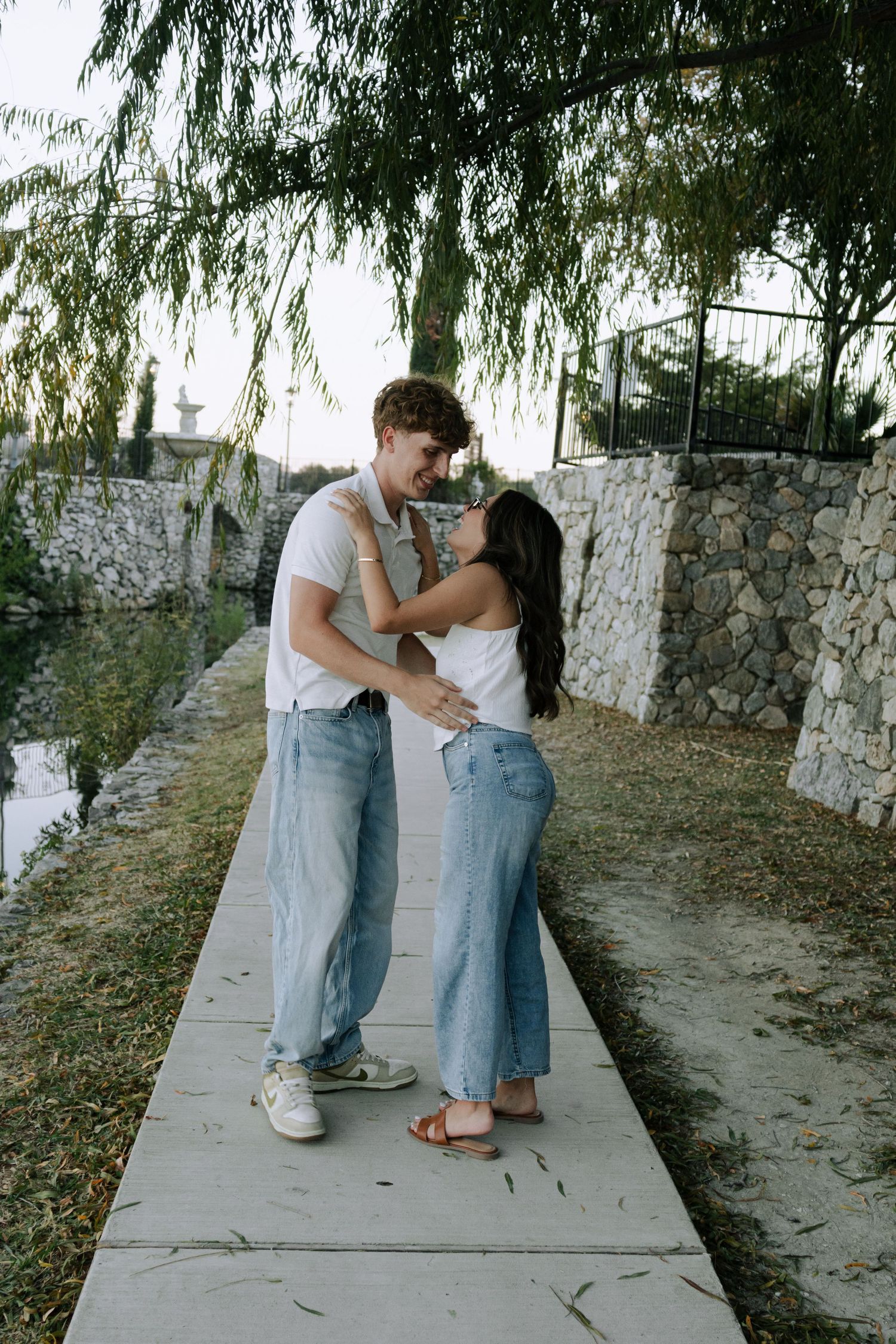 A couple in casual denim outfits share a tender moment together on a stone path under willow trees.