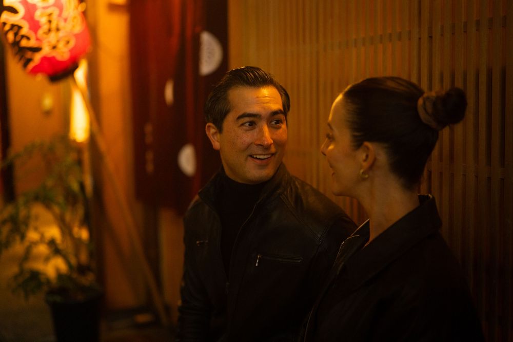 A man and woman share an intimate conversation outside a warmly lit Japanese restaurant at night.