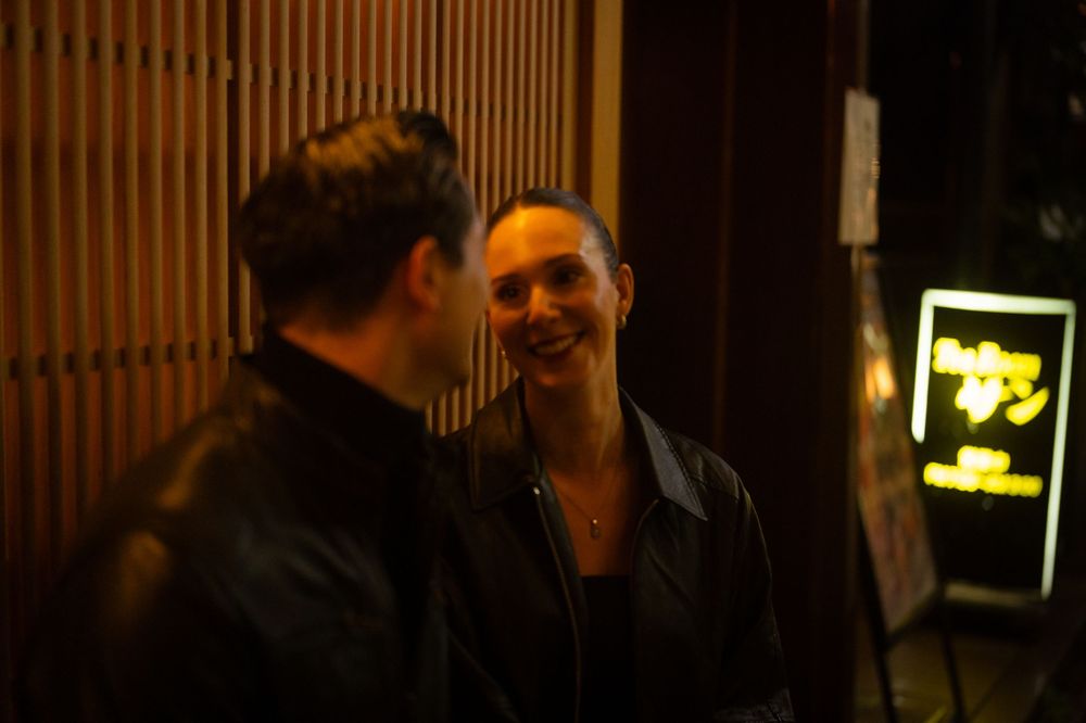 A couple smiles and laughs together outside a dimly lit bar at night.