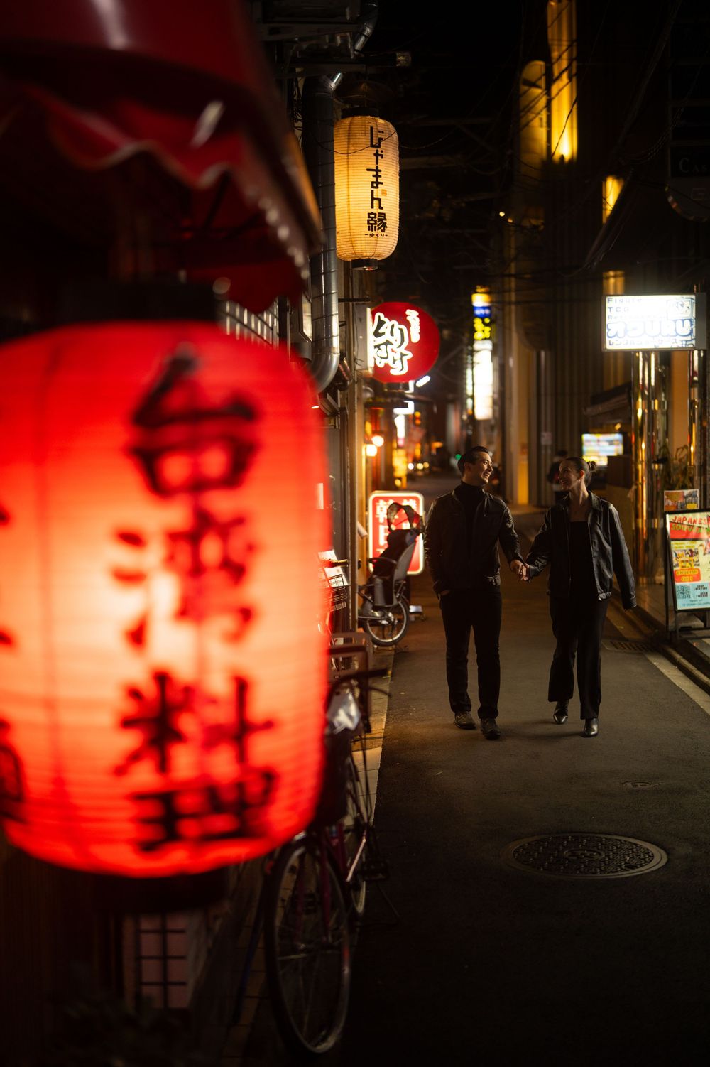 A couple walks hand in hand down a narrow Japanese alley lit by red lanterns and neon signs.