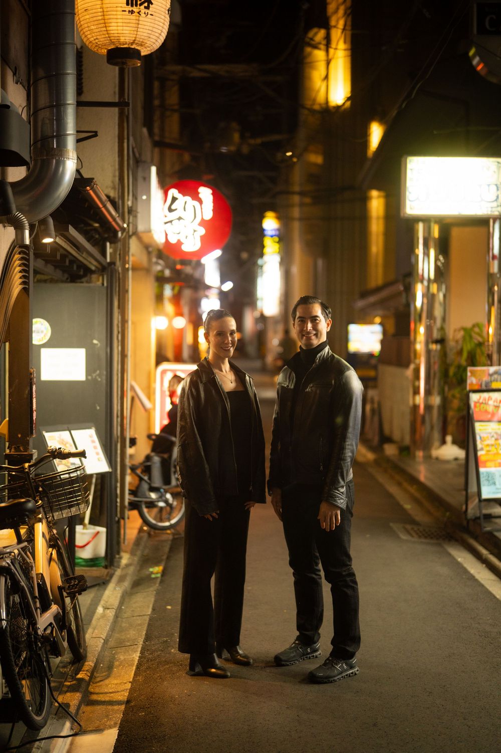 A couple poses together on a quiet Japanese street at night surrounded by glowing restaurant signs.