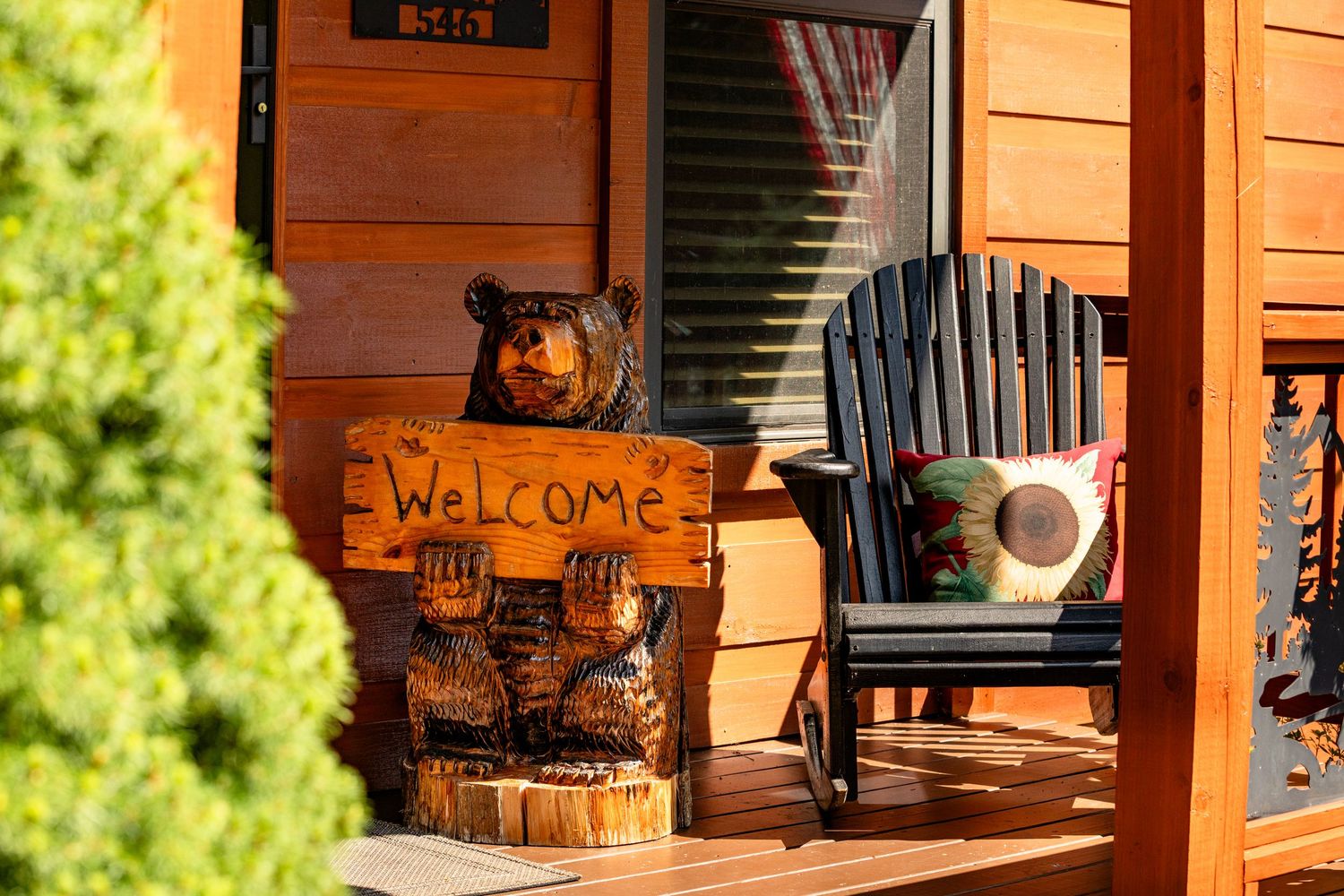 The front porch of a cabin with a carved wooden bear holding a welcome sign beside a black rocking chair with a sunflower pillow