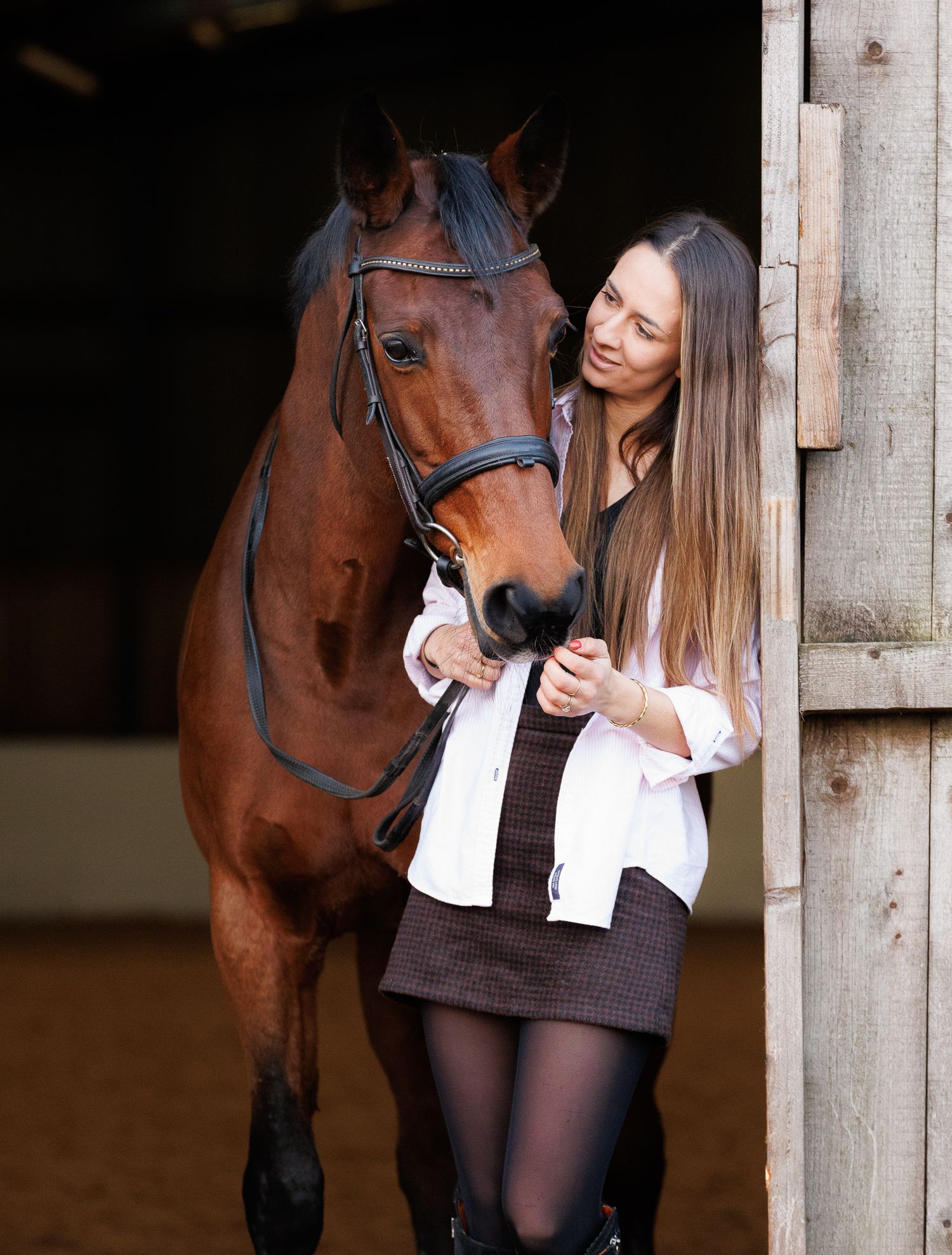 A brown horse and rider in a white sweater pose together against autumn foliage in soft natural lighting.