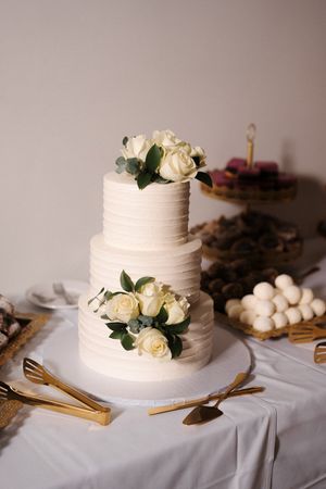 White wedding cake with textured buttercream frosting and fresh roses displayed on white tablecloth with gold utensils.