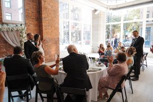 Wedding guests seated in rows at indoor ceremony venue with exposed brick walls and natural lighting.