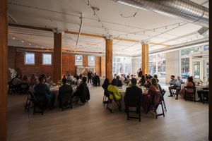 A spacious industrial-style event space filled with guests dining at round tables under string lights and exposed ductwork.